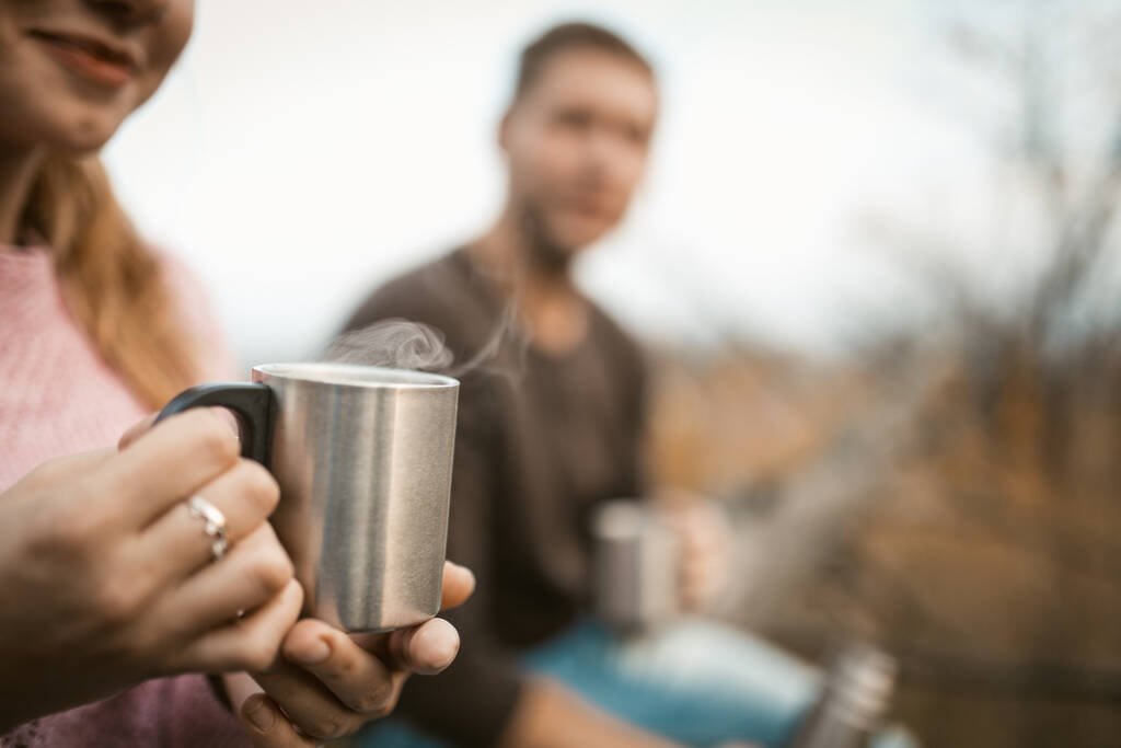 Two tourists drink a hot drink in nature. Selective focus on metal camping cup with steam from a hot coffee or tea in female hands. Caucasian guy with smiling girl have a rest outdoors. Close up shot - Photo, Image