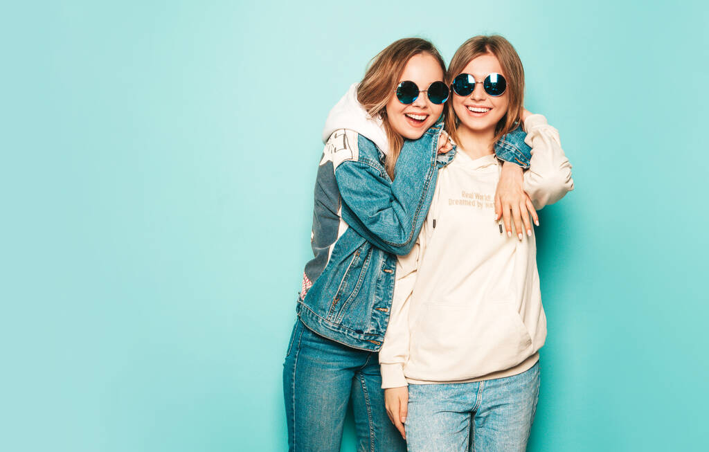 Дві молоді красиві брюнетки посміхаються дівчатам-хіпстерам у модному літньому одязі з капюшоном та джинсовою курткою. Сексуальні безтурботні жінки позують біля синьої стіни. Модні та позитивні моделі розважаються в сонцезахисних окулярах
 - Фото, зображення