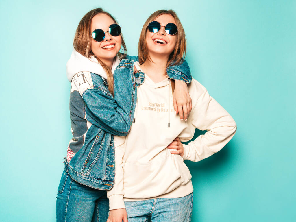 Deux jeunes belles brunes souriantes hipster girls en sweat à capuche d'été à la mode et des vêtements de veste en jean. Sexy femmes insouciantes posant près du mur bleu. Modèles tendance et positifs s'amuser dans les lunettes de soleil - Photo, image