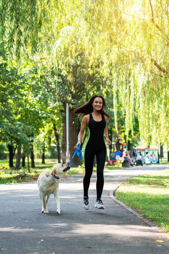 Χαρούμενη όμορφη νεαρή γυναίκα που περπατάει και τρέχει με το σκύλο της στο καλοκαιρινό πάρκο. Ενεργός ελεύθερος χρόνος, όμορφη μελαχρινή κοπέλα το πρωί τζόκινγκ με κουτάβι - Φωτογραφία, εικόνα