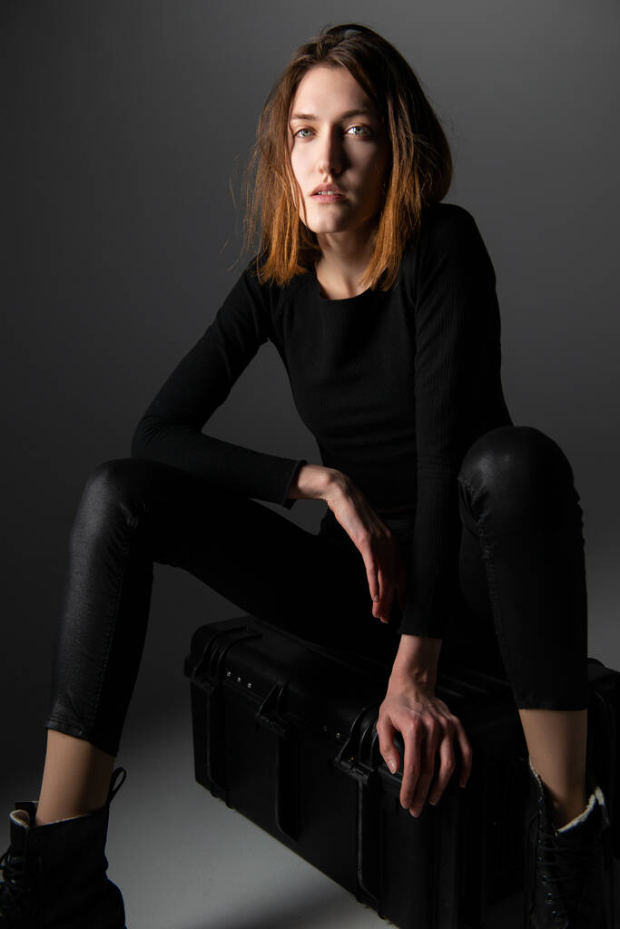 Modelo de moda. Mujer joven posando en estudio vestida de negro con botas. Hermosa chica caucásica sobre fondo gris - Foto, Imagen