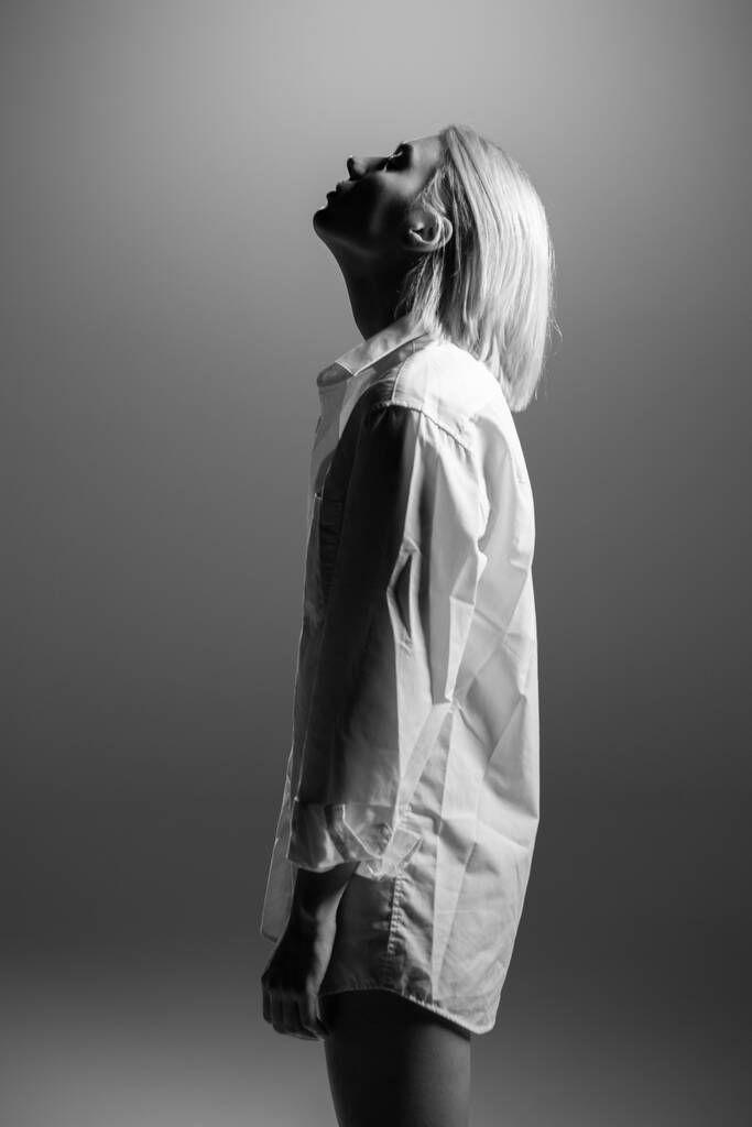 Módní model. Mladá blondýnka pózuje ve studiu v bílé košili. Krásná běloška přes šedé pozadí. Černá a bílá - Fotografie, Obrázek