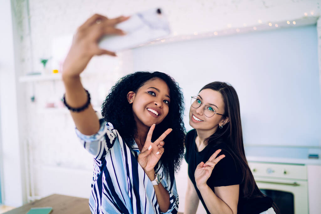 Gioioso giovane multietnico compagni di sesso femminile in abbigliamento casual mostrando segno di pace e scattare selfie in cucina moderna con interni minimalisti
 - Foto, immagini
