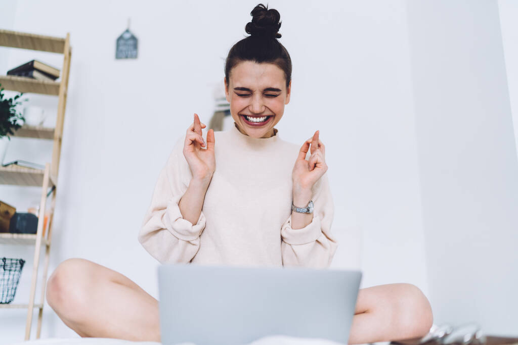 Випадкова молода жінка в вуличному одязі з великою посмішкою сидить з схрещеними ногами і пальцями і закритими очима з ноутбуком в білій кімнаті лофт
  - Фото, зображення