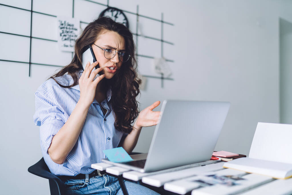 Ανήσυχη νεαρή καστανή γυναίκα με casual ρούχα επίλυση προβλημάτων μιλώντας στο κινητό και χρησιμοποιώντας laptop multitasking στο γραφείο  - Φωτογραφία, εικόνα