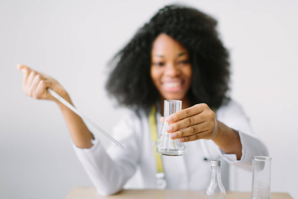 Młoda piękna Afroamerykanka lekarz w białym płaszczu ze stetoskopem. siedząc przy stole i analizując próbkę w laboratorium asystent laboratoryjny analizujący próbkę. - Zdjęcie, obraz