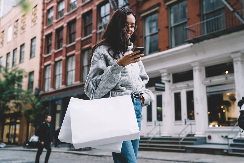 Glückliche schlanke attraktive Frau in Brille mit Einkaufstaschen in Jeans und Pullover, die Musik mit Kopfhörern per Smartphone auf der Straße hört  - Foto, Bild