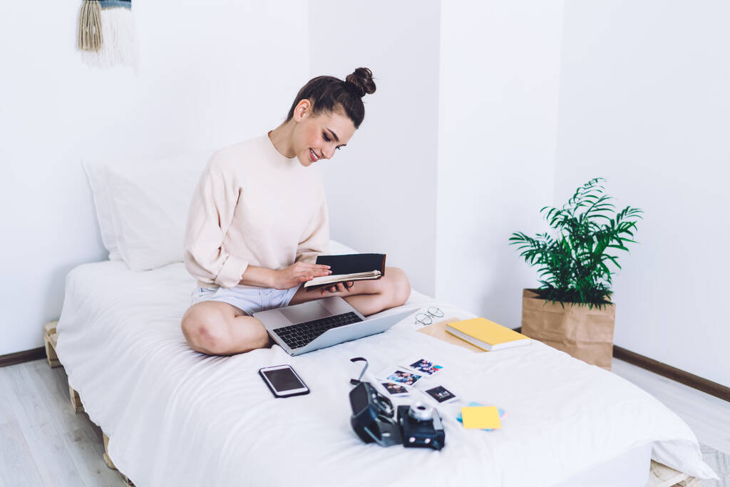 Vonzó mosolygós felnőtt nő alkalmi ruhák összpontosítva napló a kezét, miközben ül keresztbe lábak laptop az ágyon dolgokat fény minimalista fehér hálószoba - Fotó, kép