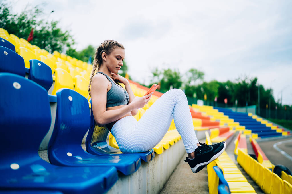 Zijaanzicht van vastberaden vrouwelijke atleet met blond haar in witte legging en grijze top zit-en surfen smartphone in het stadion - Foto, afbeelding