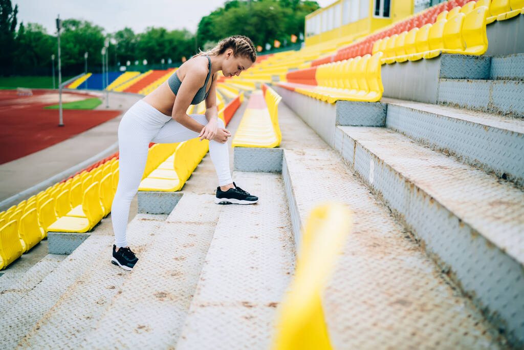 Боковой вид спортивно сфокусированной женщины с косичками, растянувшимися и отдыхающими, стоя на лестнице на трибуне, смотрящей вниз на стадион
 - Фото, изображение