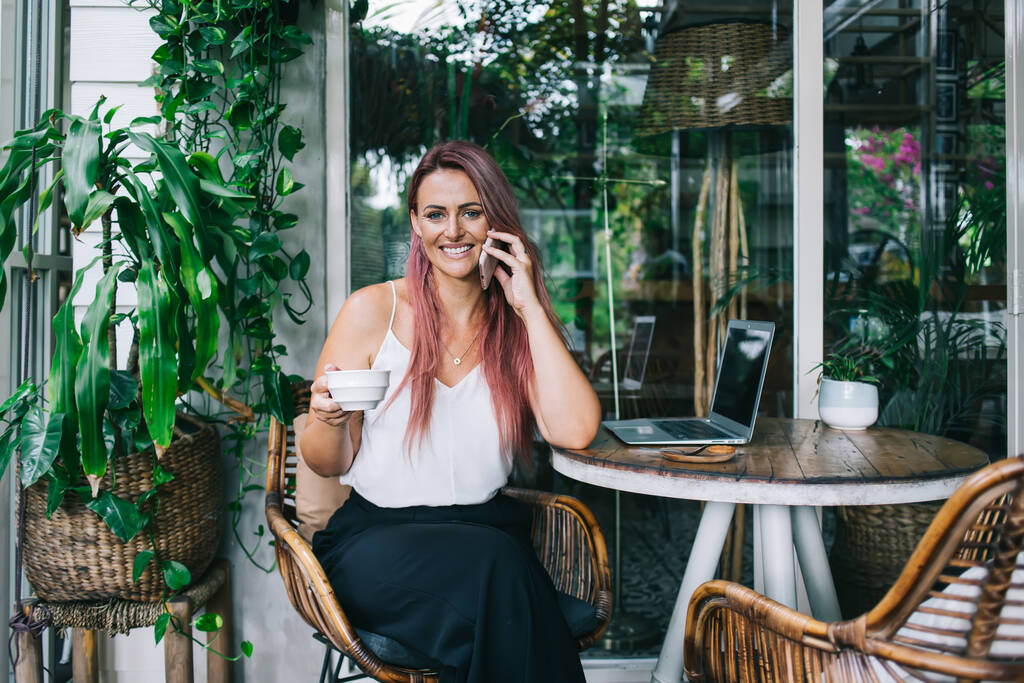 Assez réussie femme souriante assis dans un café ouvert entouré de plantes vertes avec tasse et ordinateur portable et parler sur smartphone
 - Photo, image