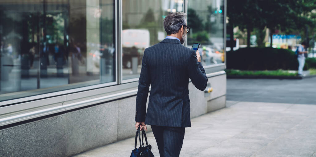 Visão traseira do empresário de cabelos grisalhos bem sucedido no elegante terno de negócios escuro moderno andando pela rua da cidade e trabalhando em smartphones em Nova York
 - Foto, Imagem