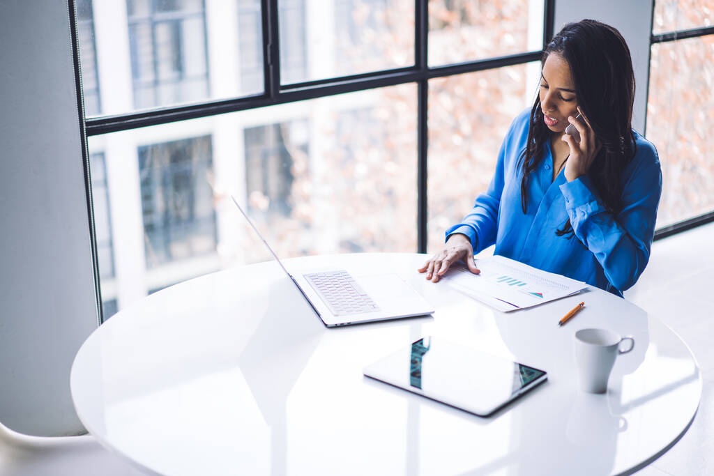 Von oben konzentrierte afroamerikanische Frau mit Laptop und Smartphone, während sie am weißen runden Tisch sitzt und Bericht in modernem Büro studiert - Foto, Bild