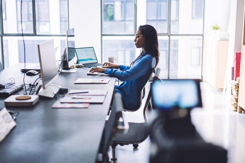 Widok z boku pewność czarny bizneswoman fotografowania na kamerze wideo procesu pracy podczas przeglądania gadżetów przy stole w nowoczesnym biurze - Zdjęcie, obraz
