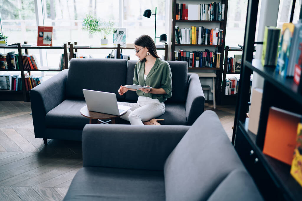 Mujer pensativa ocupada con ropa moderna sentada en un sofá acogedor mientras usa la computadora portátil y estudia documentos en la biblioteca
 - Foto, imagen
