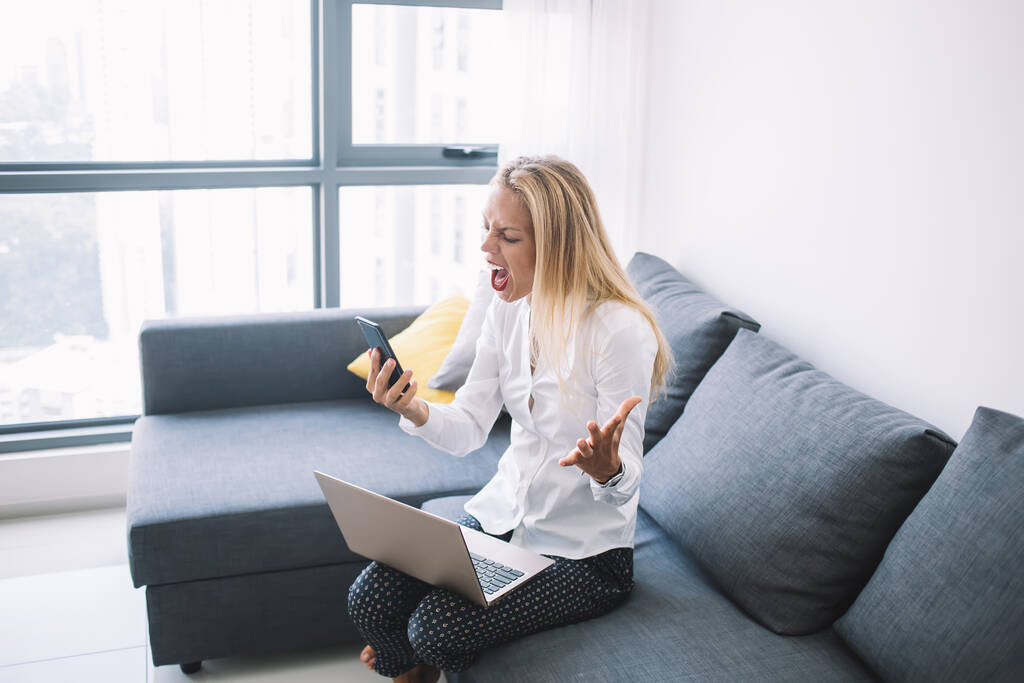 Sivunäkymä nuoresta naispuolisesta työntekijästä älypuhelimella ja vaaleilla hiuksilla istuu kannettava tietokone polvillaan sohvalla ja huutaa nostaessaan kädet toimistossa
 - Valokuva, kuva