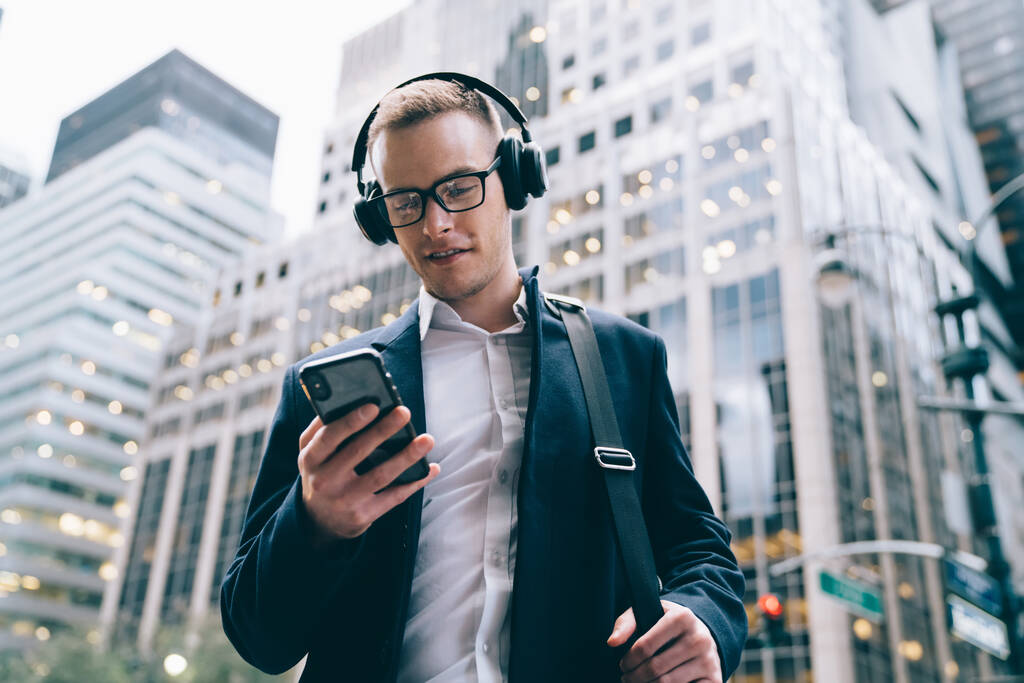 From below cheerful formal handsome male worker in glasses wearing suit holding bag and listening to music by wireless headphone while browsing mobile on blurred background of commercial buildings  - Photo, Image