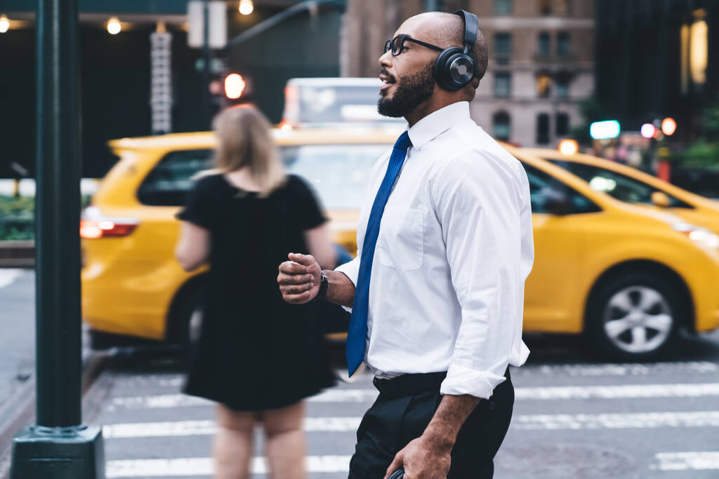 Вид сбоку на элегантного лысого и бородатого чернокожего бизнесмена в рубашке и очках в беспроводных наушниках, наслаждающегося музыкой во время прогулки на фоне размытого многолюдного центра города, глядя в сторону  - Фото, изображение