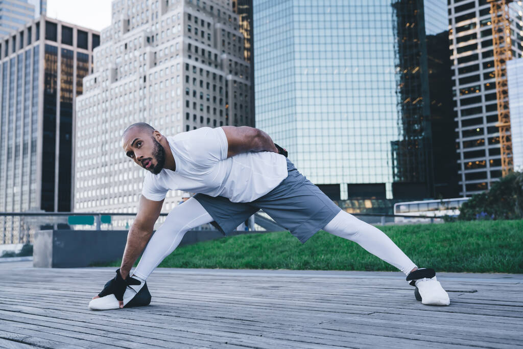 Adatto a un uomo afroamericano ben costruito in abbigliamento sportivo che allunga le gambe sul marciapiede di legno di fronte a grattacieli al mattino guardando altrove - Foto, immagini