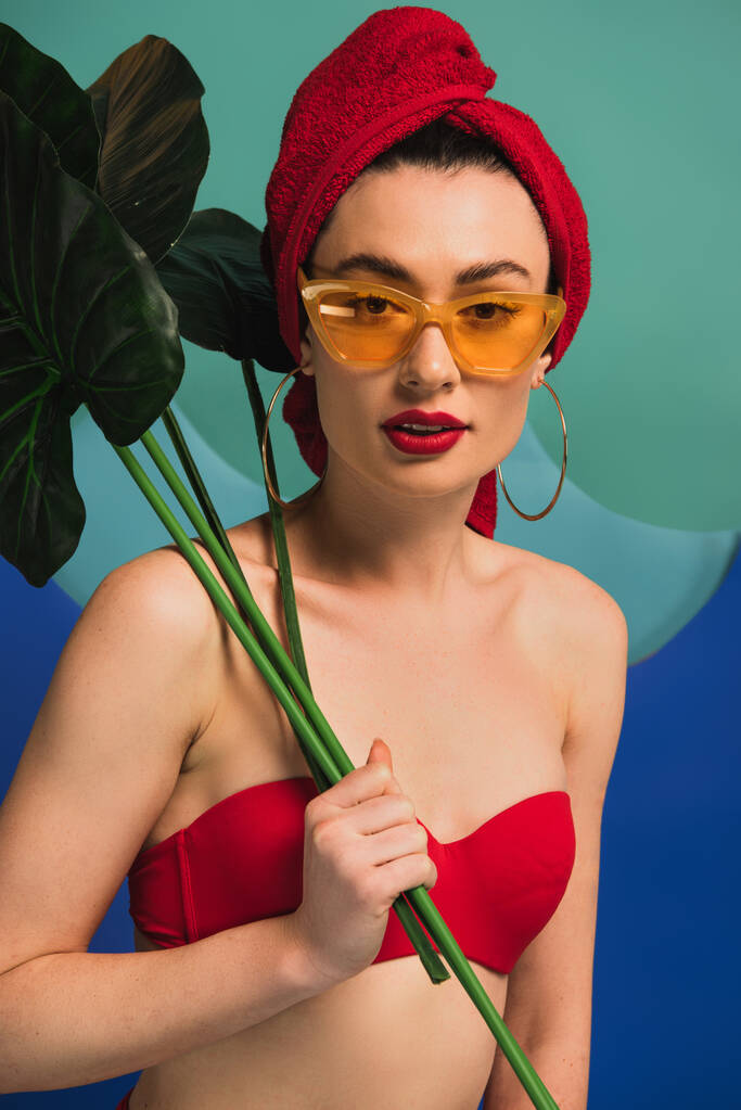ελκυστικό κορίτσι με κόκκινη πετσέτα μαγιό και γυαλιά ηλίου κρατώντας τροπικά φύλλα φοίνικα σε μπλε και τυρκουάζ - Φωτογραφία, εικόνα