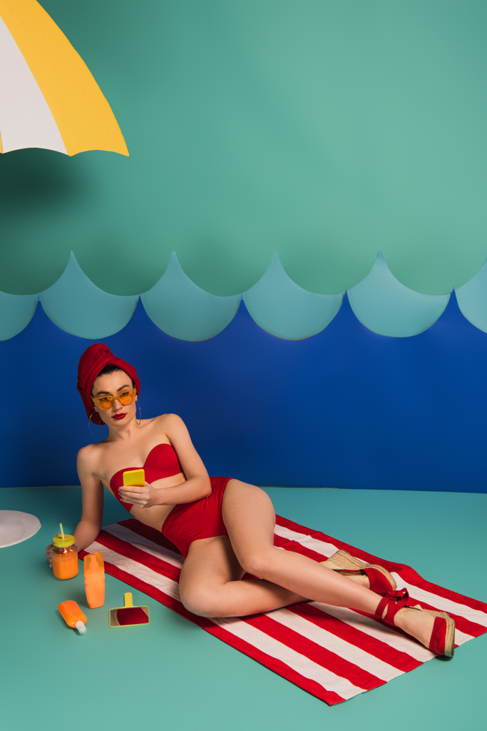 Kırmızı mayolu ve güneş gözlüklü çekici bir kadın kağıt kesiği dalgalarının yanında akıllı telefon kullanıyor.  - Fotoğraf, Görsel