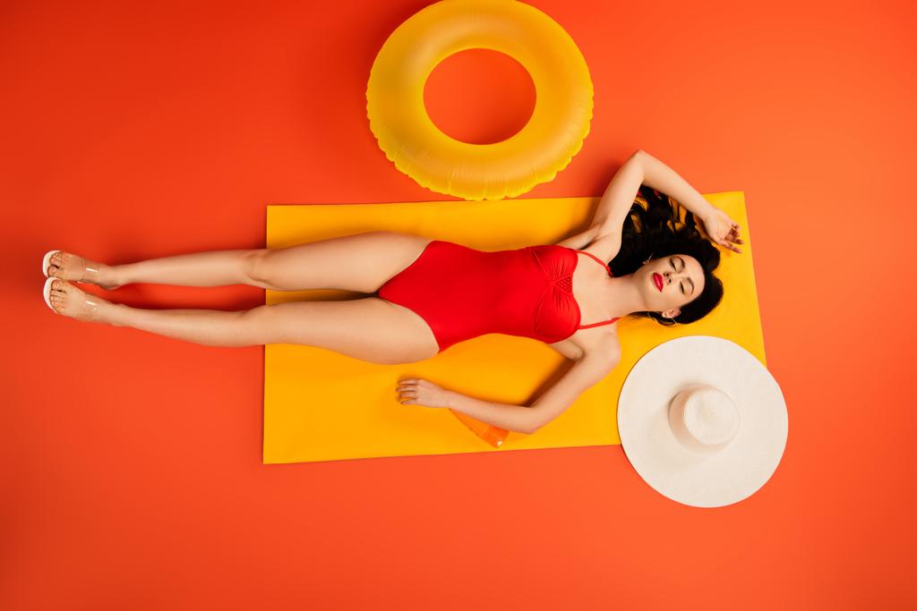 top view of girl in bathing suit lying near bottle with sunscreen, inflatable ring, straw hat and mirror on orange - Photo, Image