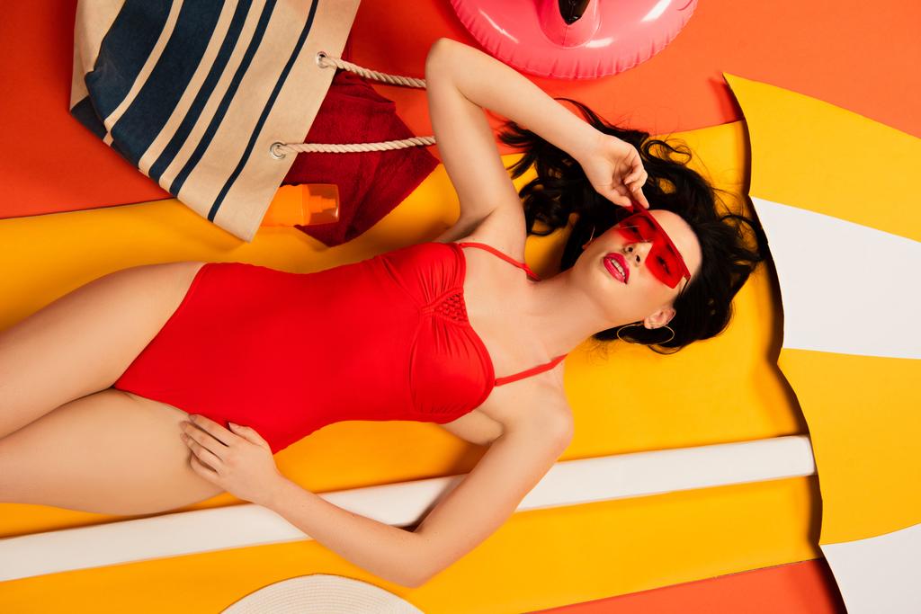 vista superior de la hermosa mujer en gafas de sol y traje de baño tocando la tapa y acostado en naranja
  - Foto, Imagen