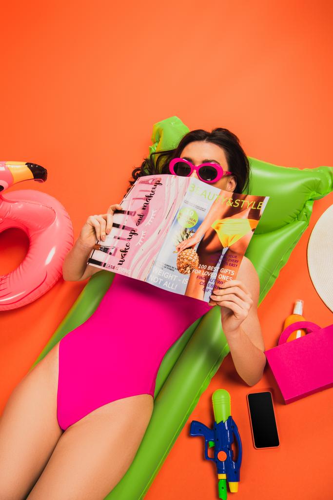 young woman in sunglasses and swimsuit lying on inflatable mattress and covering face with magazine on orange - Photo, Image
