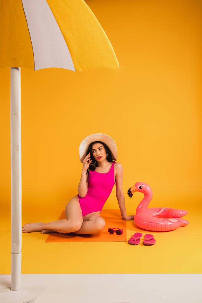 mujer joven en sombrero de paja y traje de baño sentado cerca de las gafas de sol, chanclas, anillo inflable y hablando en el teléfono inteligente en amarillo
 - Foto, imagen