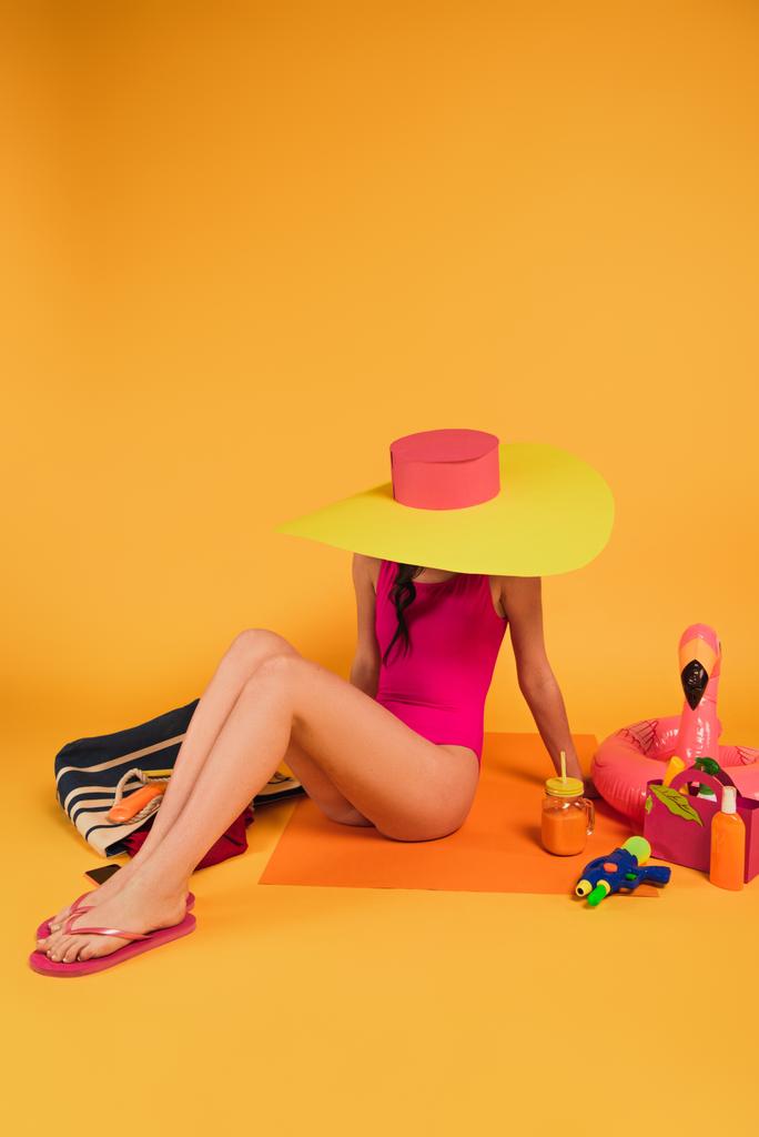 elegante ragazza in cappello di paglia e costume da bagno rosa seduta vicino al vetro con succo d'arancia, pistola ad acqua e anello gonfiabile su giallo
 - Foto, immagini