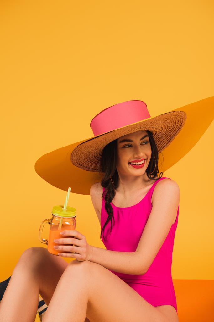 alegre chica en sombrero de paja y traje de baño celebración de vidrio con jugo de naranja en amarillo
 - Foto, imagen