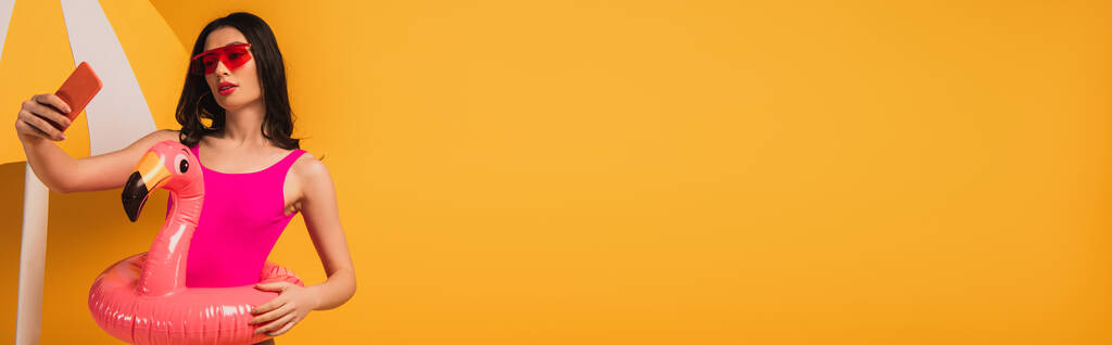 imagen horizontal de chica atractiva en gafas de sol y traje de baño de pie con anillo inflable y tomar selfie en amarillo
  - Foto, imagen