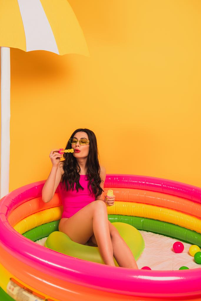 young woman in swimsuit blowing soap bubbles while sitting in inflatable pool on yellow  - Photo, Image