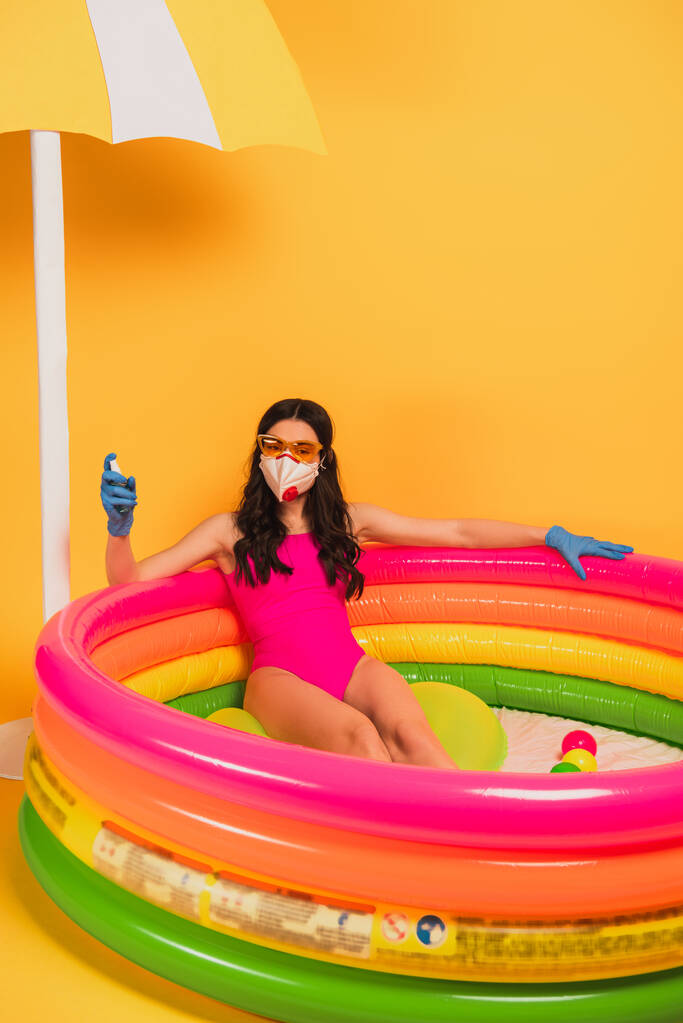 junge Frau in Badeanzug, Sonnenbrille, Latexhandschuhen und medizinischer Maske sitzt im aufblasbaren Pool und hält Desinfektionsmittel auf gelb - Foto, Bild