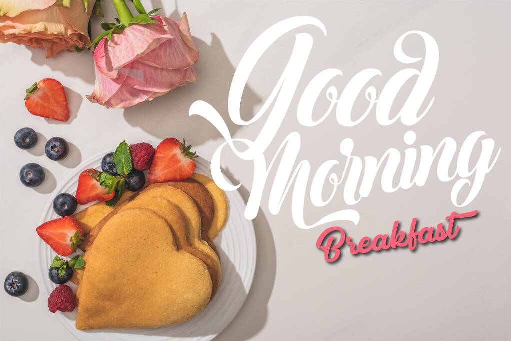 Draufsicht auf köstliche herzförmige Pfannkuchen mit Beeren in der Nähe von Rosen auf weißem Hintergrund, guten Morgen Illustration - Foto, Bild