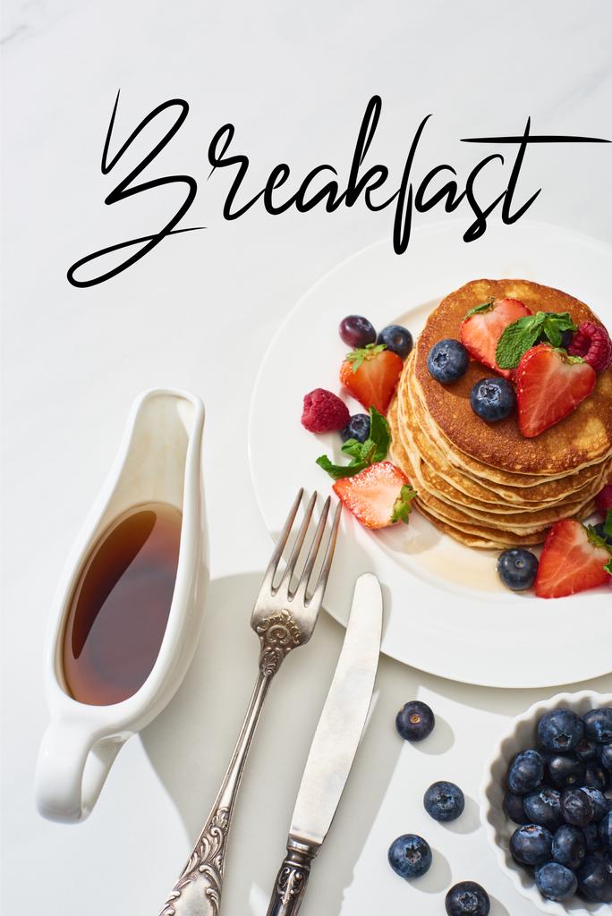 top view of delicious pancakes with maple syrup, blueberries and strawberries on plate near fork and knife on marble white surface, breakfast illustration - Photo, Image