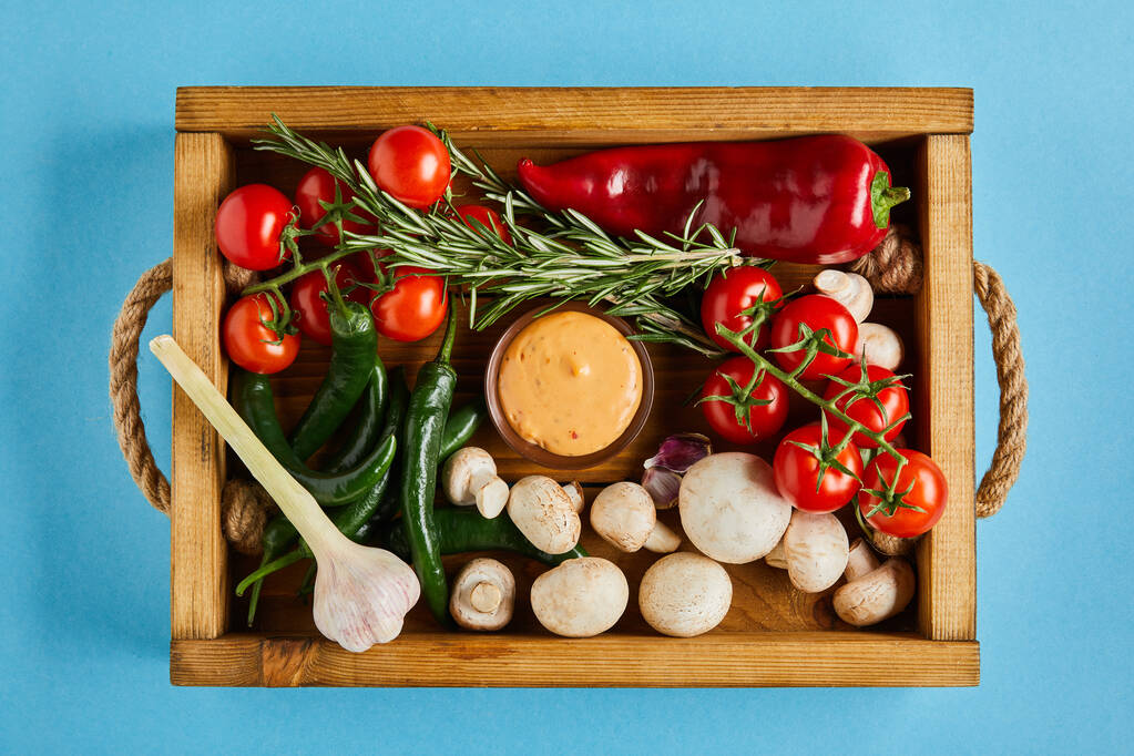 vrchní pohled na lahodnou omáčku v misce v blízkosti čerstvé zralé zeleniny, rozmarýnu a hub v dřevěné krabici na modrém pozadí - Fotografie, Obrázek