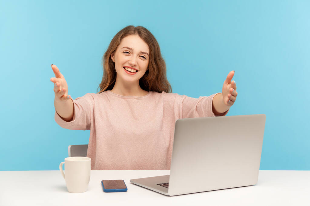 Vítej, příteli! Šťastný příjemný mladý žena zaměstnanec sedí na pracovišti s notebookem a natahuje ruce, aby objal, pozdravuje klienta s úsměvem. vnitřní studio záběr izolované na modrém pozadí - Fotografie, Obrázek
