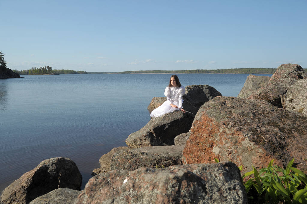 jong meisje in een witte jurk zit in de buurt van het water op de stenen - Foto, afbeelding