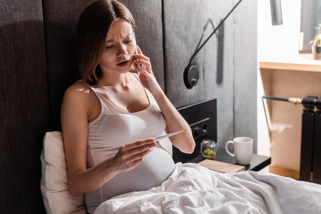 mujer embarazada joven hablando en el teléfono inteligente y mirando termómetro digital en el dormitorio
  - Foto, Imagen