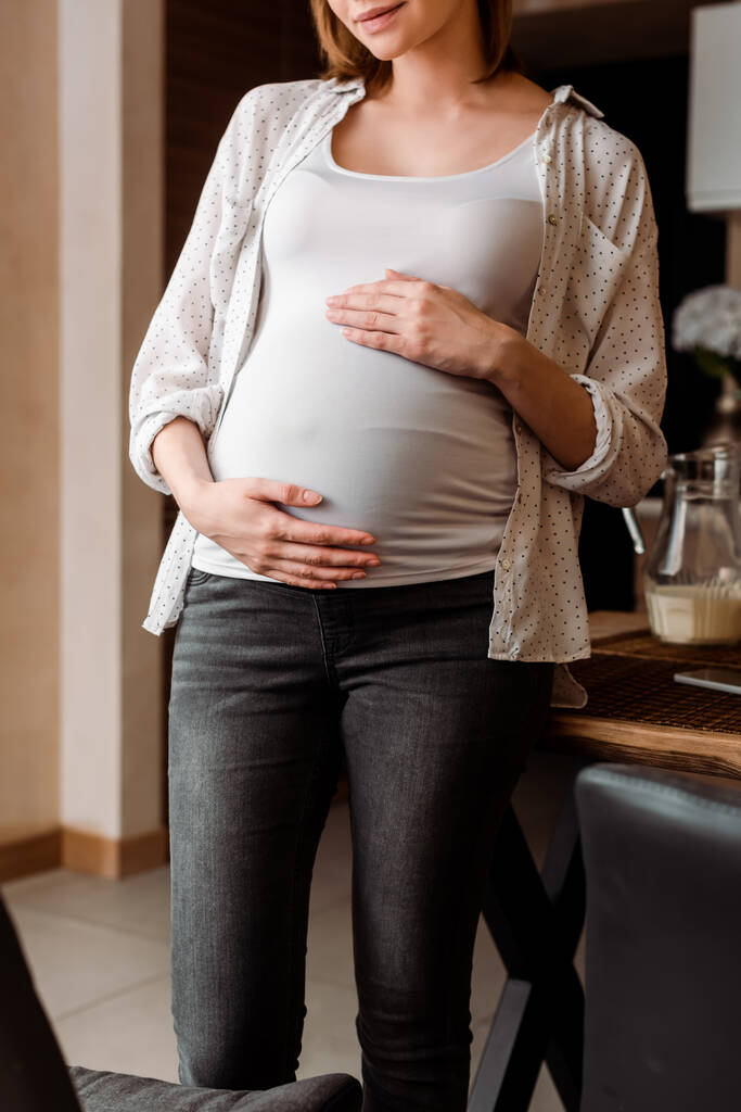 abgeschnittene Ansicht einer schwangeren Frau, die ihren Bauch berührt, während sie zu Hause steht - Foto, Bild