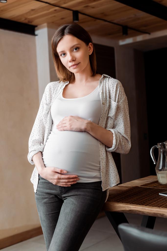 mujer atractiva y embarazada tocando el vientre mientras está de pie en casa
 - Foto, Imagen