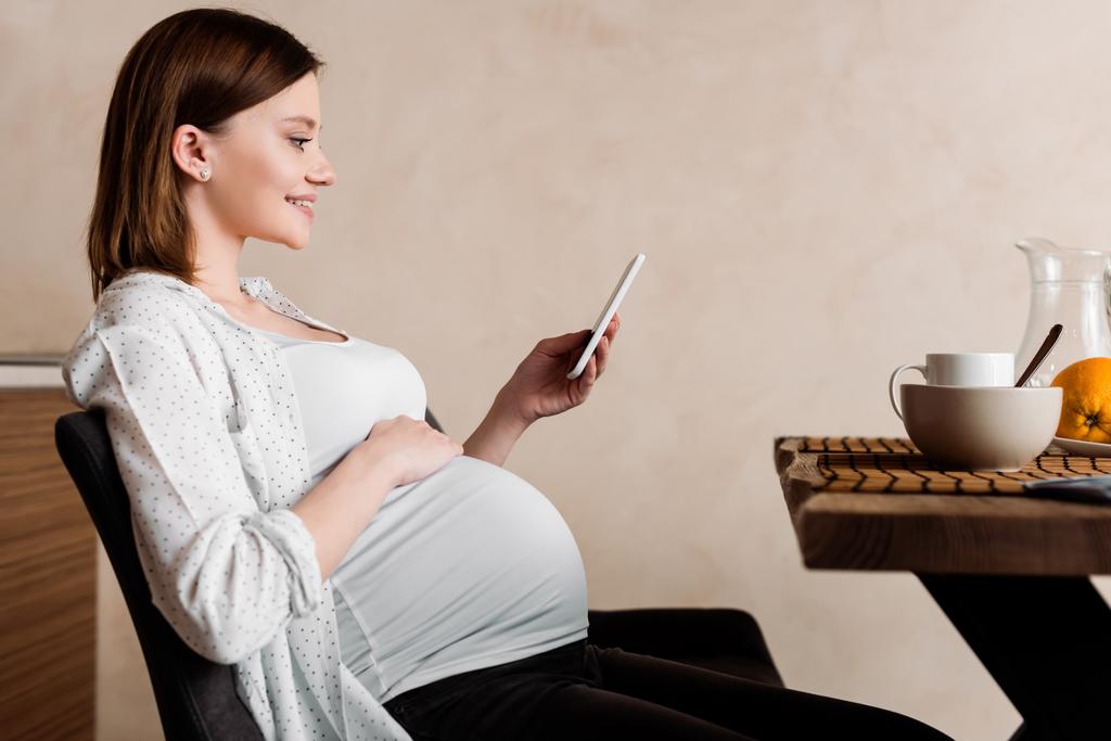 vista lateral de la mujer embarazada feliz utilizando el teléfono inteligente cerca del desayuno sabroso
  - Foto, Imagen