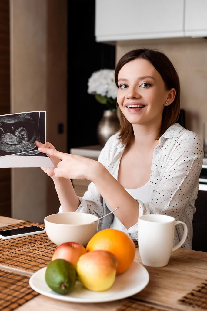 selective focus of cheerful and pregnant woman pointing with finger at ultrasound photos near fruits and smartphone with blank screen - Photo, Image
