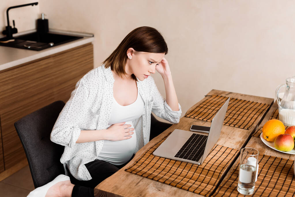 Schwangere Freiberuflerin berührt Bauch in der Nähe von Laptop, Smartphone, Glas Wasser und Obst auf dem Tisch - Foto, Bild