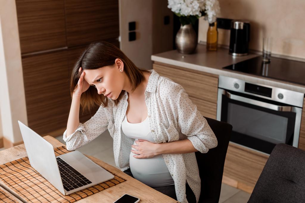 uupunut ja raskaana freelancer käyttäen kannettavaa tietokonetta koskettaessaan vatsa kotona
 - Valokuva, kuva