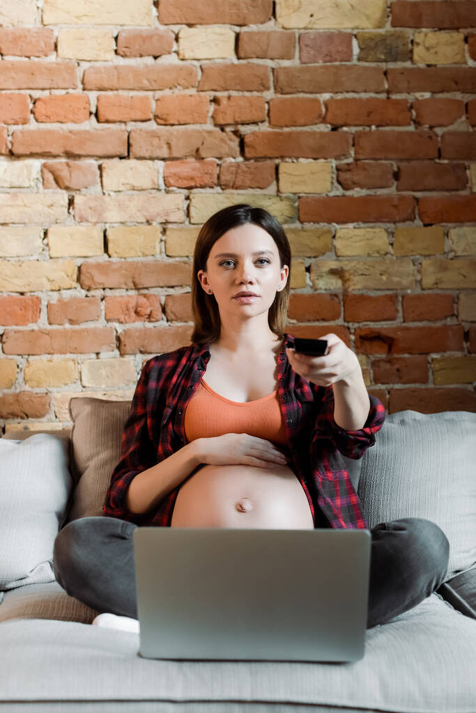 zwanger vrouw met afstandsbediening en het kijken naar film in de buurt laptop  - Foto, afbeelding