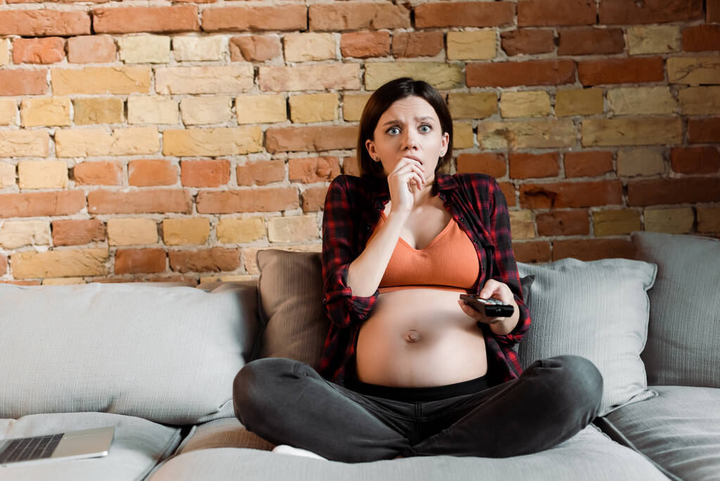 scared and pregnant woman holding remote controller and watching movie - Photo, Image