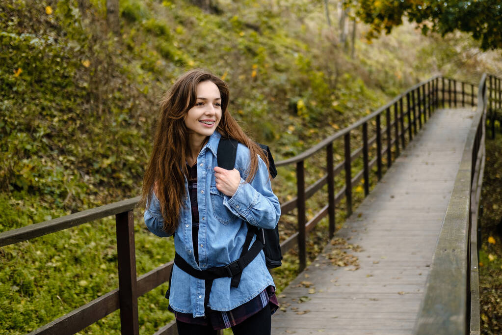 Kobieta turystka z plecakiem spaceruje po moście w jesiennym lesie. - Zdjęcie, obraz
