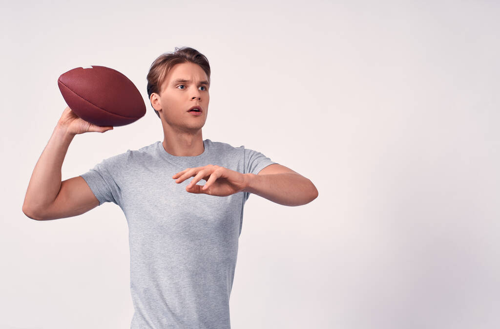 Mladý atletický fitness atletický běloch s blonďatými vlasy, v šedém základním tričku, drží v pravých rukou americký fotbalový míček, připravený hodit. Kopírovat prostor. - Fotografie, Obrázek